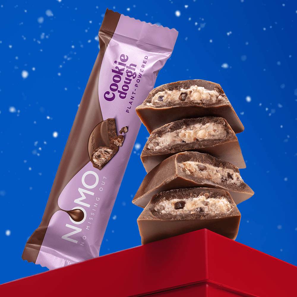 A package of NOMO Cookie Dough plant-based chocolate bar stands next to a stack of three sliced chocolate-coated cookies filled with cookie dough, set against a blue snowy background and a red box corner.