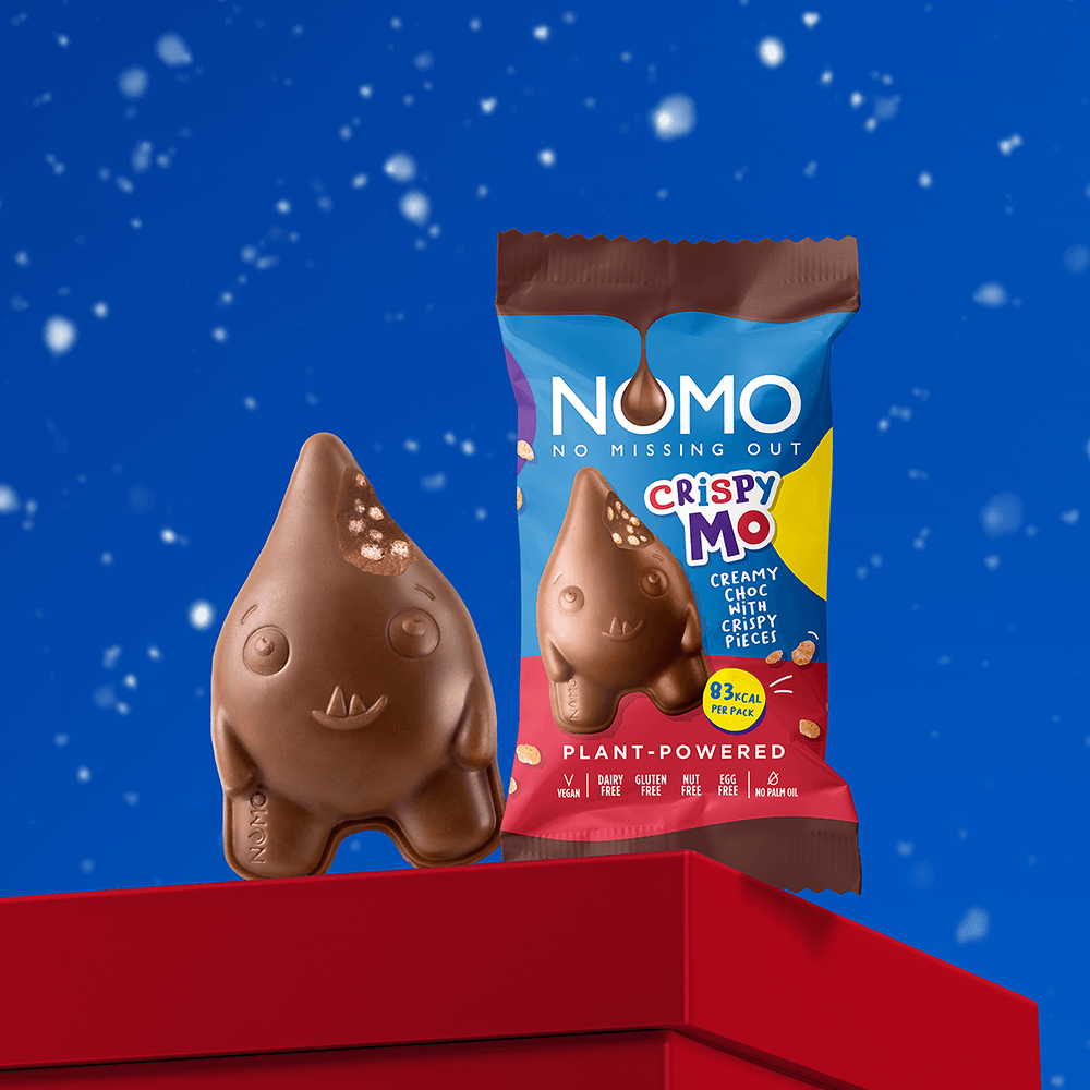 A chocolate monster-shaped NOMO Crispy Mo piece stands next to its branded bag on a red platform against a blue snowy background. 